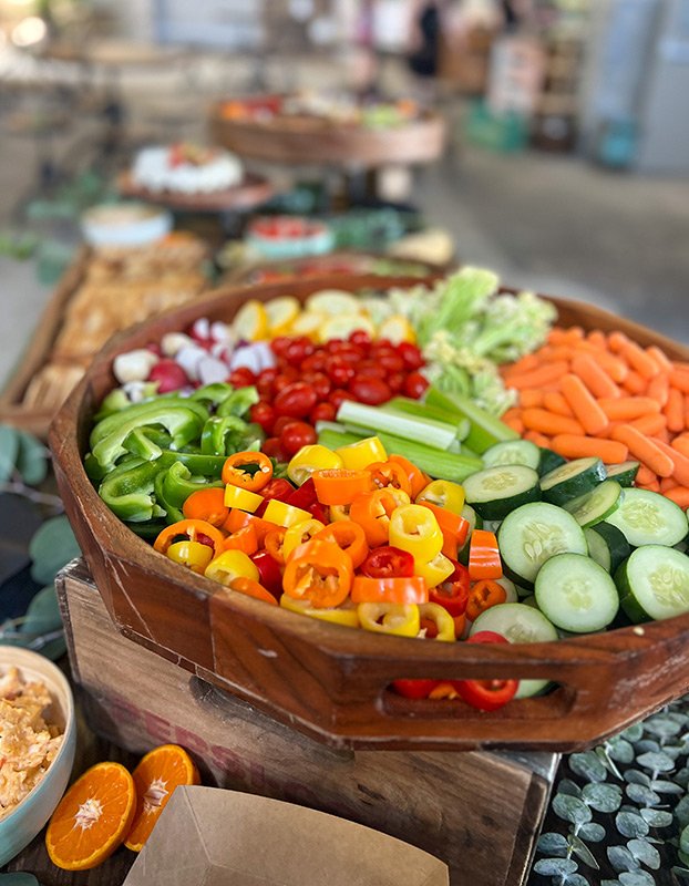 Colorful sliced vegetables including peppers, cherry tomatoes, cucumbers, celery, and carrots in a wooden bowl at a market.