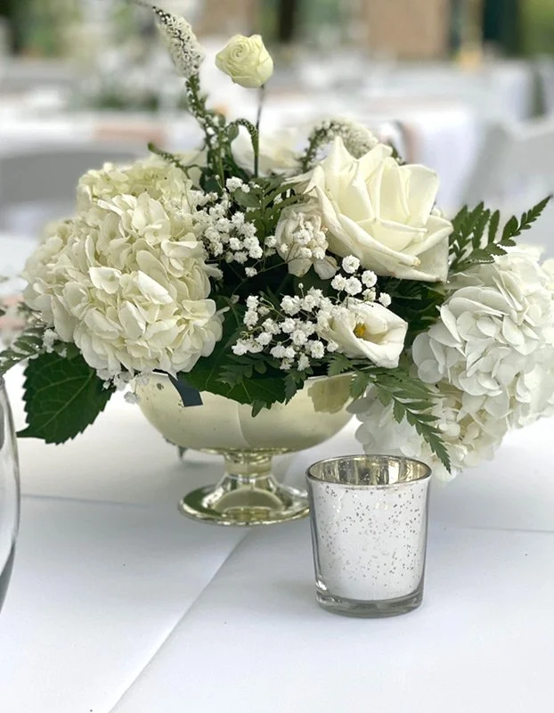 A floral centerpiece with white roses, hydrangeas, and baby's breath in a silver bowl, placed on a white tablecloth with a small silver candle holder nearby.
