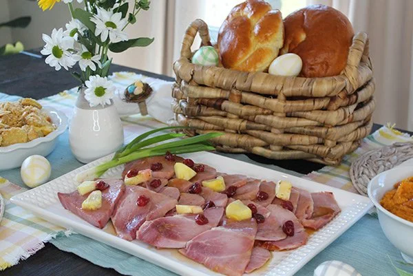 Easter breakfast table with sliced ham, pineapple chunks, cranberries, decorated eggs, bread rolls in a basket, daisies in a vase, and side dishes.