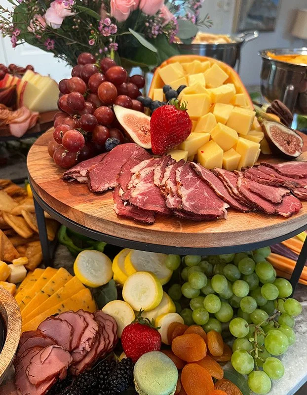 A tiered cheese and charcuterie platter featuring cubes of cheese, sliced cured meats, red and green grapes, strawberries, figs, and assorted fruits, garnished with flowers in the background.