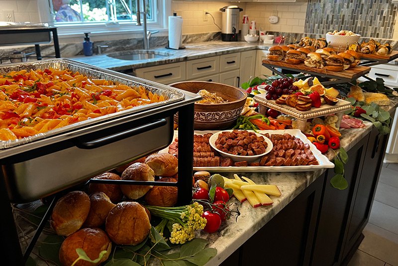 A breakfast buffet with a baked pasta dish, bread rolls, sliced meats, cheeses, fresh vegetables, fruits, and pastries on a kitchen counter.