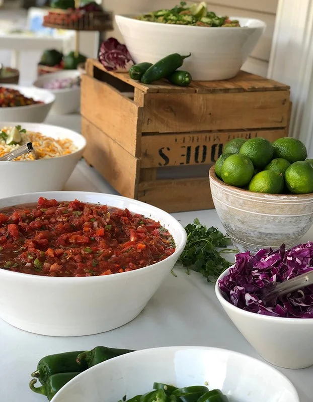 A variety of fresh vegetables and salads on a table, including limes, jalapenos, purple cabbage, and bowls of salsa and shredded vegetables, with a large bowl of salad on a wooden crate in the background.