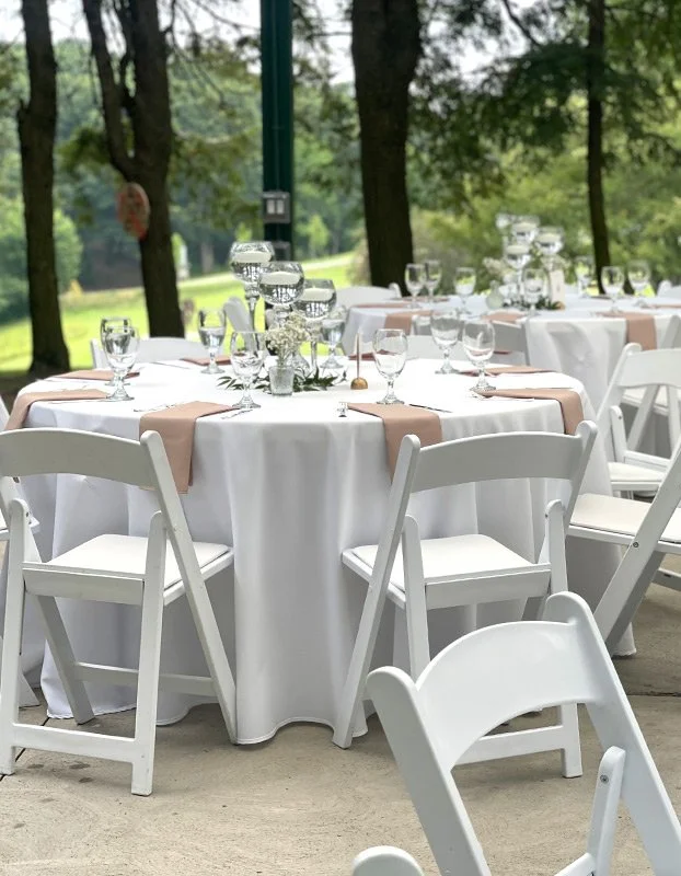 Outdoor event setup with round tables covered in white tablecloths, roses and candle centerpieces, glassware, and white chairs amid trees.