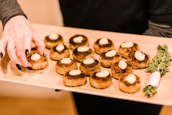 Person holding a wooden board with several croquettes topped with white sauce and a sprig of greenery on the side.
