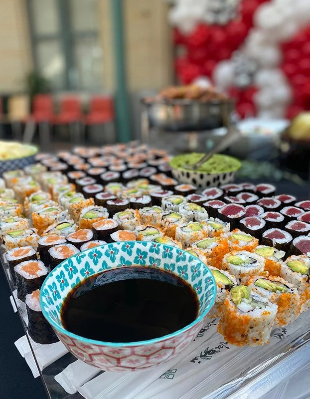 Various types of sushi rolls arranged on a table, with a bowl of soy sauce in the foreground and a blurred background with more food and red and white decorations.