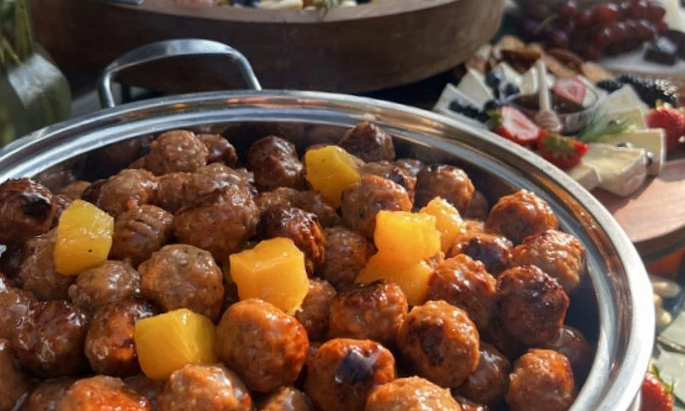 A large glass dish filled with cooked meatballs topped with chunks of pineapple, with a background of a cheese platter with strawberries, grapes, and various cheeses.