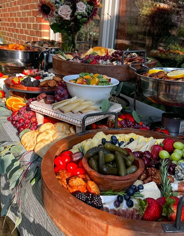 A lavish outdoor charcuterie spread on a table, featuring various cheeses, fruits, vegetables, crackers, and a large flower bouquet in the background.