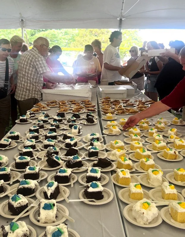 Table filled with cupcakes and cakes at a crowded outdoor event under a tent, with people serving and selecting desserts.