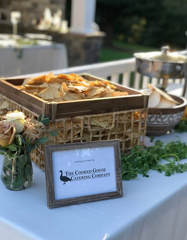 Tray of sliced bread on a table at an outdoor catering event with a sign reading 'Catering provided by The Cooked Goose Catering Company' and a flower arrangement in a small vase.