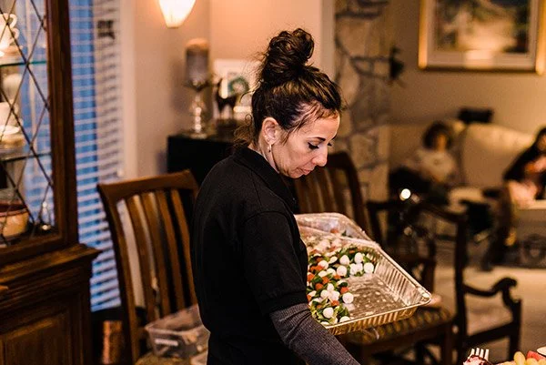 A woman with dark hair tied back in a bun looks down at a gold-colored tray filled with small white and pink decorations inside a living room.