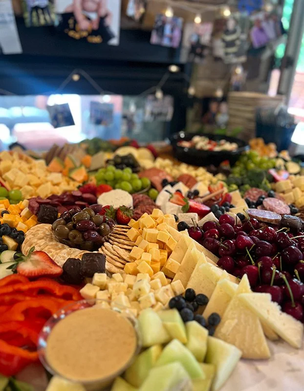 A large cheese platter with various cheeses, grapes, strawberries, cherries, honey, and sliced vegetables at a party or event.