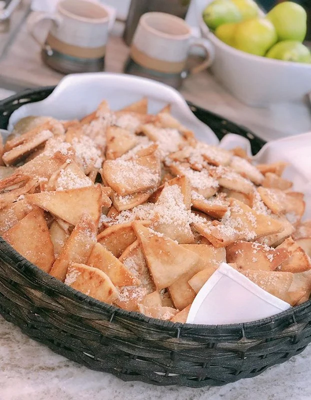 Basket of fried wonton chips sprinkled with grated cheese or salt, with coffee mugs and a bowl of green apples in the background.