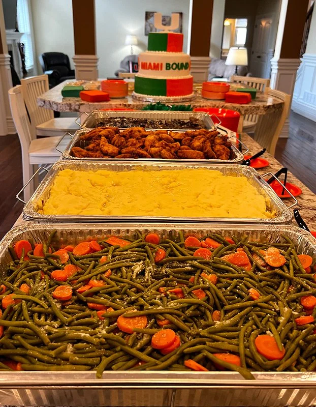 Buffet table with green bean casserole, mashed potatoes, fried chicken, and a decorated cake in the background with a 'Miami Bound' message.