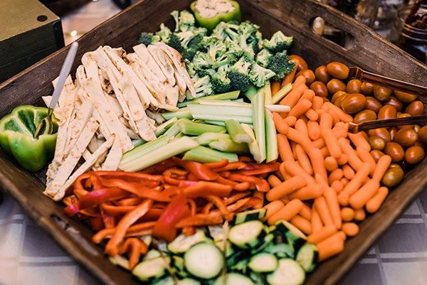A wooden tray filled with assorted fresh vegetables including broccoli, celery, grape tomatoes, baby carrots, red bell peppers, cucumber slices, green bell pepper, and sliced chicken strips.