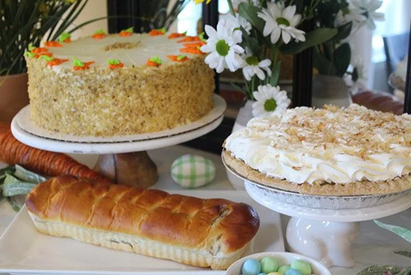A table with a cake decorated with flowers, a cream cake with coconut flakes, a bread roll, and a bowl of colorful candy eggs, with flowers in the background.