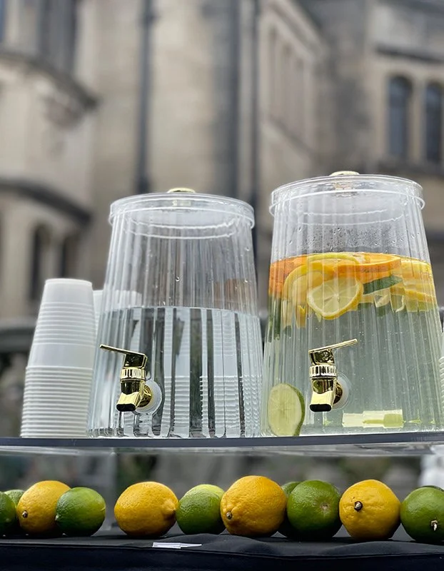 Water dispenser with lemon and orange slices, clear plastic cups, and fresh lemons and limes on a tray outside.