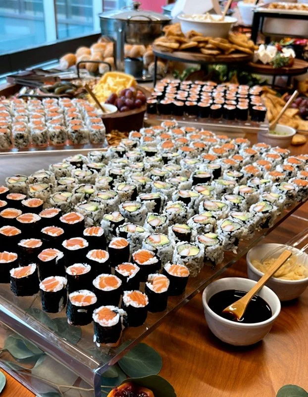 Assorted sushi rolls arranged on a large transparent tray, with soy sauce, wasabi, and ginger on the side, at a buffet table with other foods in the background.