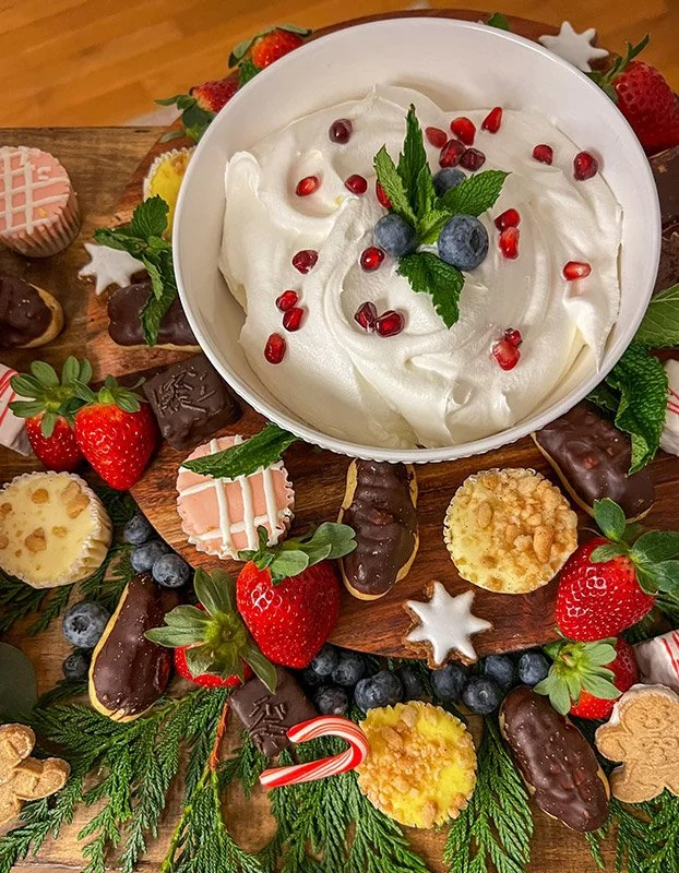 A festive Christmas dessert platter with a bowl of whipped cream topped with pomegranate seeds, blueberries, and mint leaves, surrounded by strawberries, assorted cookies, berries, and holiday decorations on a wooden board with greenery.