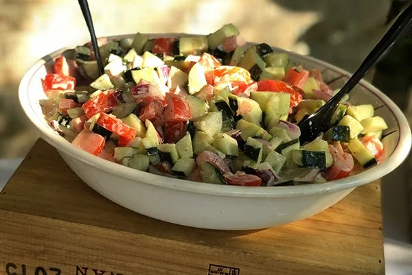 A bowl of chopped vegetable salad with cucumbers, tomatoes, and olives, with serving utensils inside.