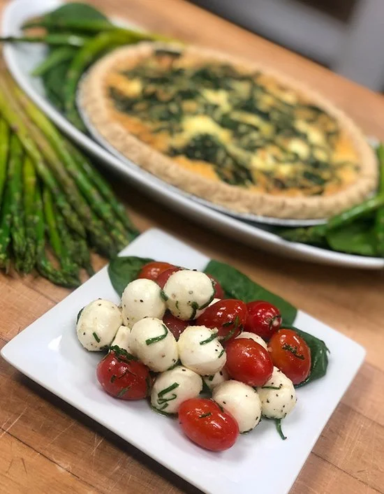 Plate of cherry tomatoes and mozzarella balls garnished with basil, with a spinach and cheese quiche and fresh asparagus on a wooden table.