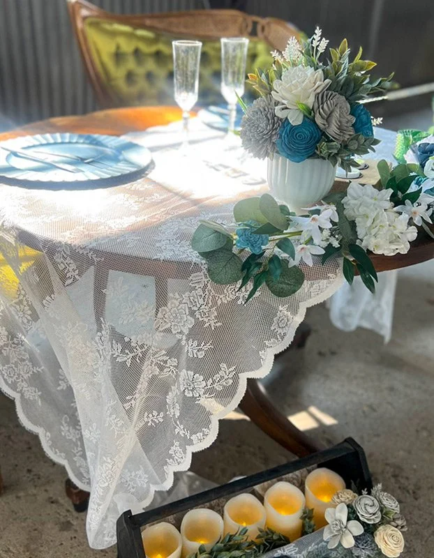 A table decorated with a white lace tablecloth, floral centerpiece, and place settings with blue plates. Two champagne glasses are set on the table. There is a row of candles in a black tray below the table, and a wooden chair is visible in the backg