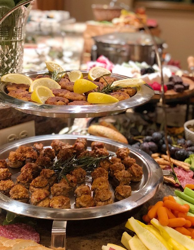 Two silver platters of fried chicken pieces garnished with herbs and lemon wedges, with a variety of other foods and condiments in the background on a buffet table.