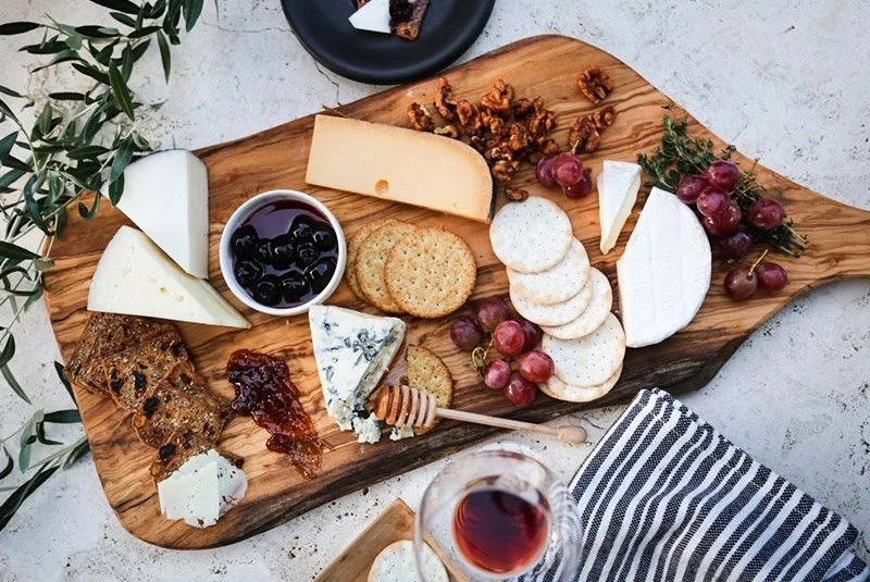 Cheese platter with various cheeses, crackers, grapes, honey, nuts, and a glass of red wine on a wooden serving board.