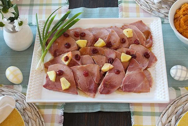 A white plate with sliced ham garnished with pineapple chunks and red berries, garnished with green onions, on a checkered tablecloth with Easter eggs nearby.