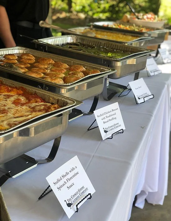 Buffet table with trays of food including stuffed chicken breasts with spinach florentine sauce, meatballs, a vegetable dish, and a cheesy casserole, with labels in front of each dish.