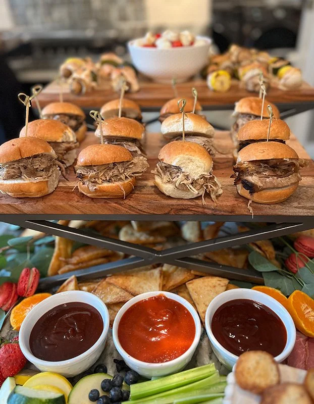 Assorted mini sandwiches with shredded meat on buns, three bowls of different sauces, and fresh fruit with strawberries, blueberries, orange slices, celery sticks, and crostini on a decorative tray.