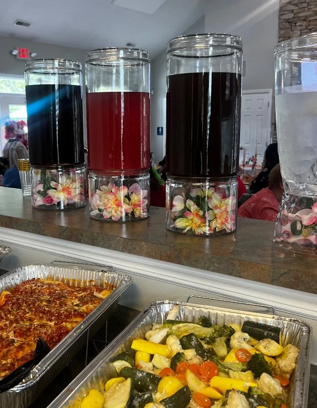 Three glass containers filled with colorful liquids (blue, red, and dark brown) on a restaurant counter with floral decorations on the bottom, alongside a glass of water, with a pizza and vegetable tray in the foreground, and a restaurant interior wi