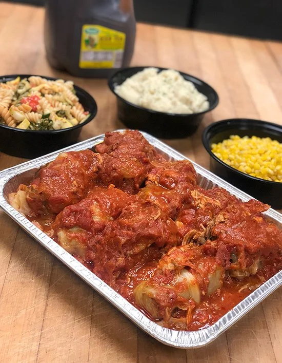 A tray of baked chicken drumsticks with tomato sauce, accompanied by bowls of white rice, pasta salad with vegetables, and yellow corn on a wooden table.