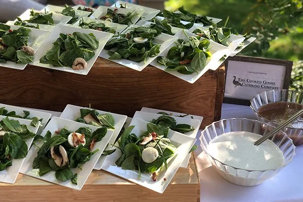 Small plates with spinach, mushrooms, cheese, and prosciutto next to bowls of white sauce or dressing, on a table outdoors.