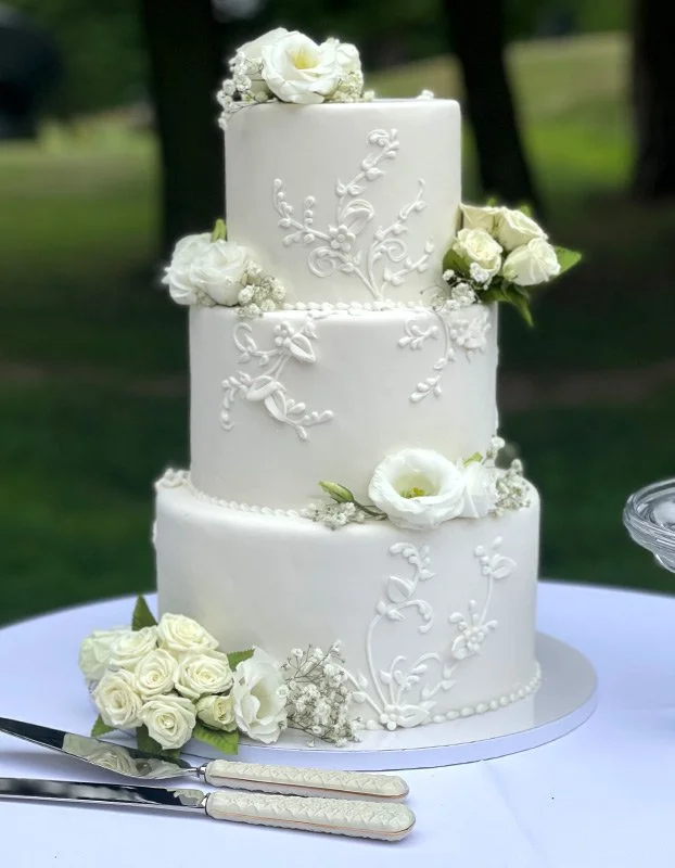 Three-layer white wedding cake with floral decorations and piping, set on a white table with wedding utensils and greenery in the background.