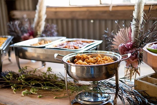 Buffet table with chicken and pizza dishes, decorated with flowers and pampas grass.
