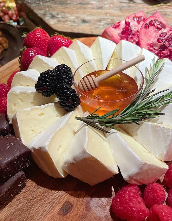 Cheese wheel garnished with blackberries, raspberries, and a sprig of rosemary, accompanied by honey in a glass bowl with a honey dipper, on a wooden surface, with strawberries and sliced pomegranate in the background.