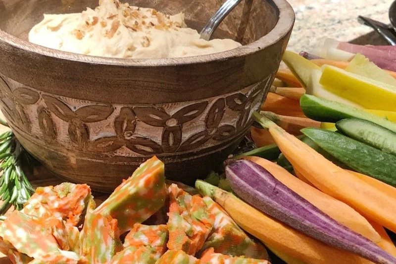 A bowl of creamy dip with a spoon, surrounded by fresh vegetables including cucumber, carrots, and zucchini, and a pile of snack crackers.