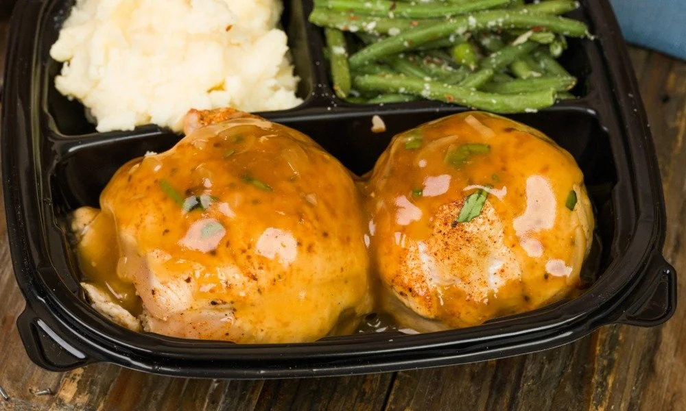 A meal in a black divided container with mashed potatoes, green beans, and two pieces of chicken covered in gravy.