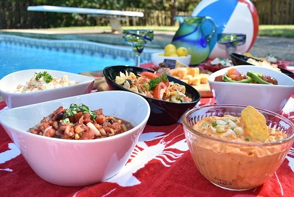 Outdoor poolside party with a red tablecloth, featuring bowls of salsa, guacamole, and dip, along with drinks, watermelon floaties, and yellow lemons.