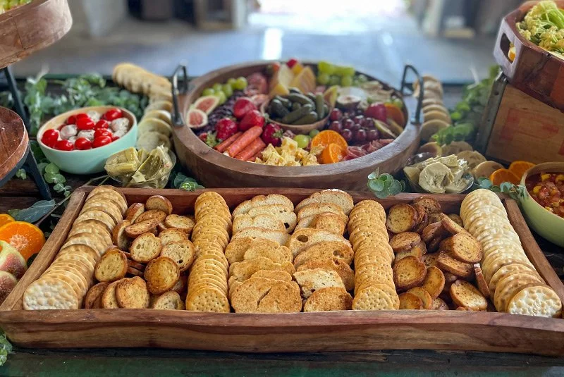 Tray of assorted crackers with dips and a bowl of cherry tomatoes, surrounded by a fruit platter and salad bowls.