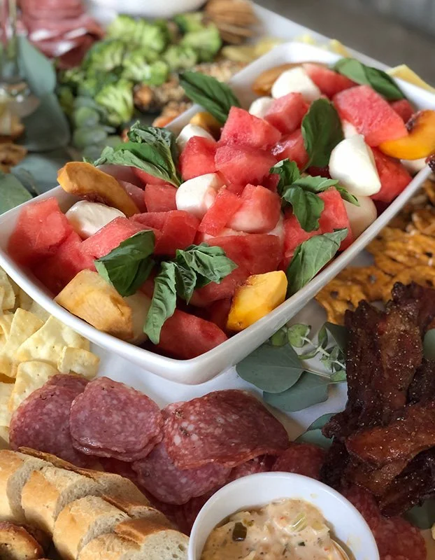 A platter with fresh fruit salad containing watermelon, mango, and mozzarella, along with sliced cured meats, bread, and barbecue ribs.