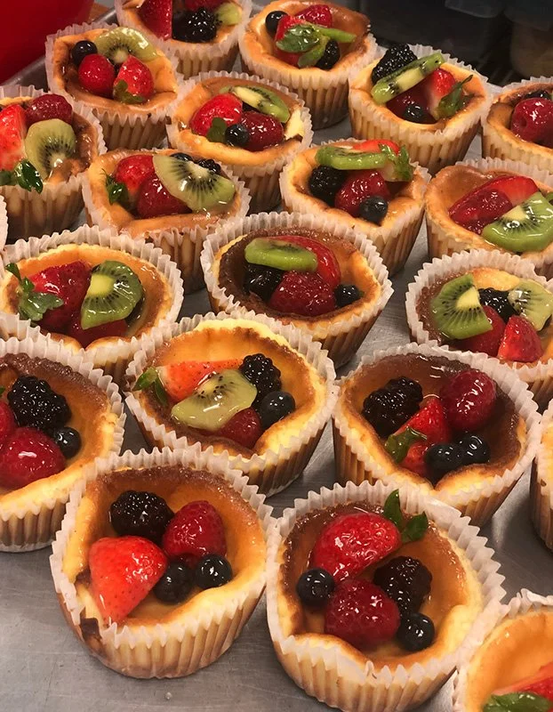 Assorted mini fruit tarts topped with strawberries, blueberries, blackberries, kiwi slices, and small green leaves in paper cups.