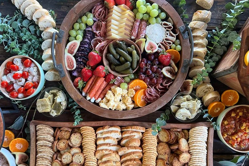 Charcuterie board with cheeses, grapes, figs, strawberries, pickles, meats, crackers, and assorted fruits, accompanied by bowls of cherry tomatoes, artichokes, and a vegetable salad, decorated with eucalyptus leaves.