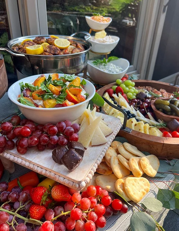 A variety of food and snacks on a table, including grapes, strawberries, cheese, crackers, a salad, fruit, and a bowl of meatballs with lemons.