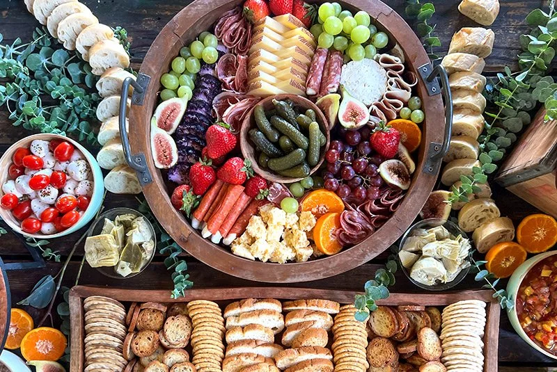 Charcuterie board with assorted meats, cheeses, grapes, strawberries, figs, pickles, and crackers. Surrounded by small bowls of cherry tomatoes, artichoke hearts, and a bowl of chopped fruit salad. Decorated with eucalyptus leaves.