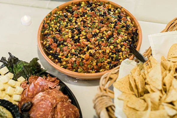 A large bowl of colorful corn and bean salad with a serving spoon, surrounded by a cheese and meat platter and a basket of tortilla chips.