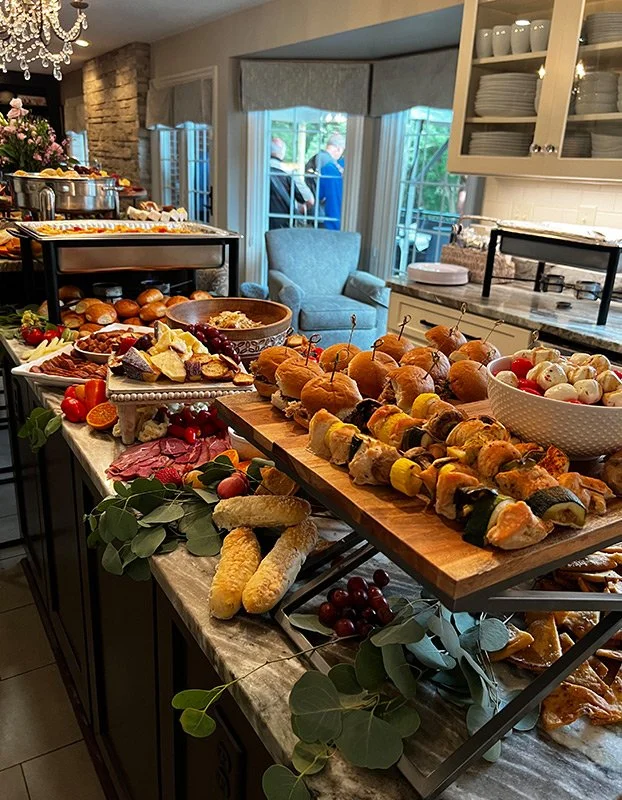 A spread of various appetizers and finger foods on a kitchen island, including skewers, rolls, cheese, meats, vegetables, and grapes, with a buffet setup in the background.