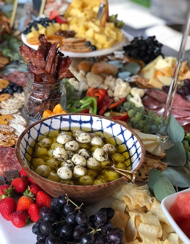Close-up of a Mediterranean-style appetizer platter featuring a bowl of marinated mozzarella balls in olive oil, fresh strawberries and black grapes, crispy bacon in a jar, sliced cheese, pineapple chunks, assorted meats, and vegetables, with additio