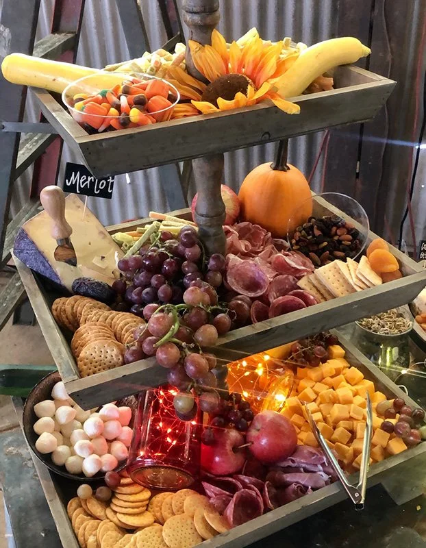 Three-tiered wooden tray table decorated with assorted cheeses, grapes, crackers, cured meats, and fruits such as bananas and sunflower, resembling a festive or party snack display.