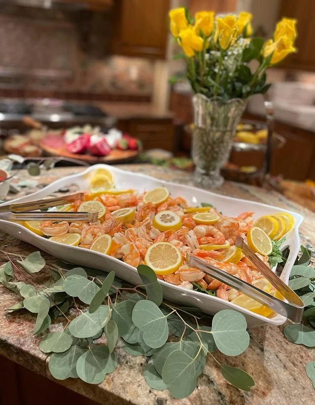 Shrimp cocktail garnished with lemon slices on a white platter, surrounded by eucalyptus leaves, with a vase of yellow roses in the background.